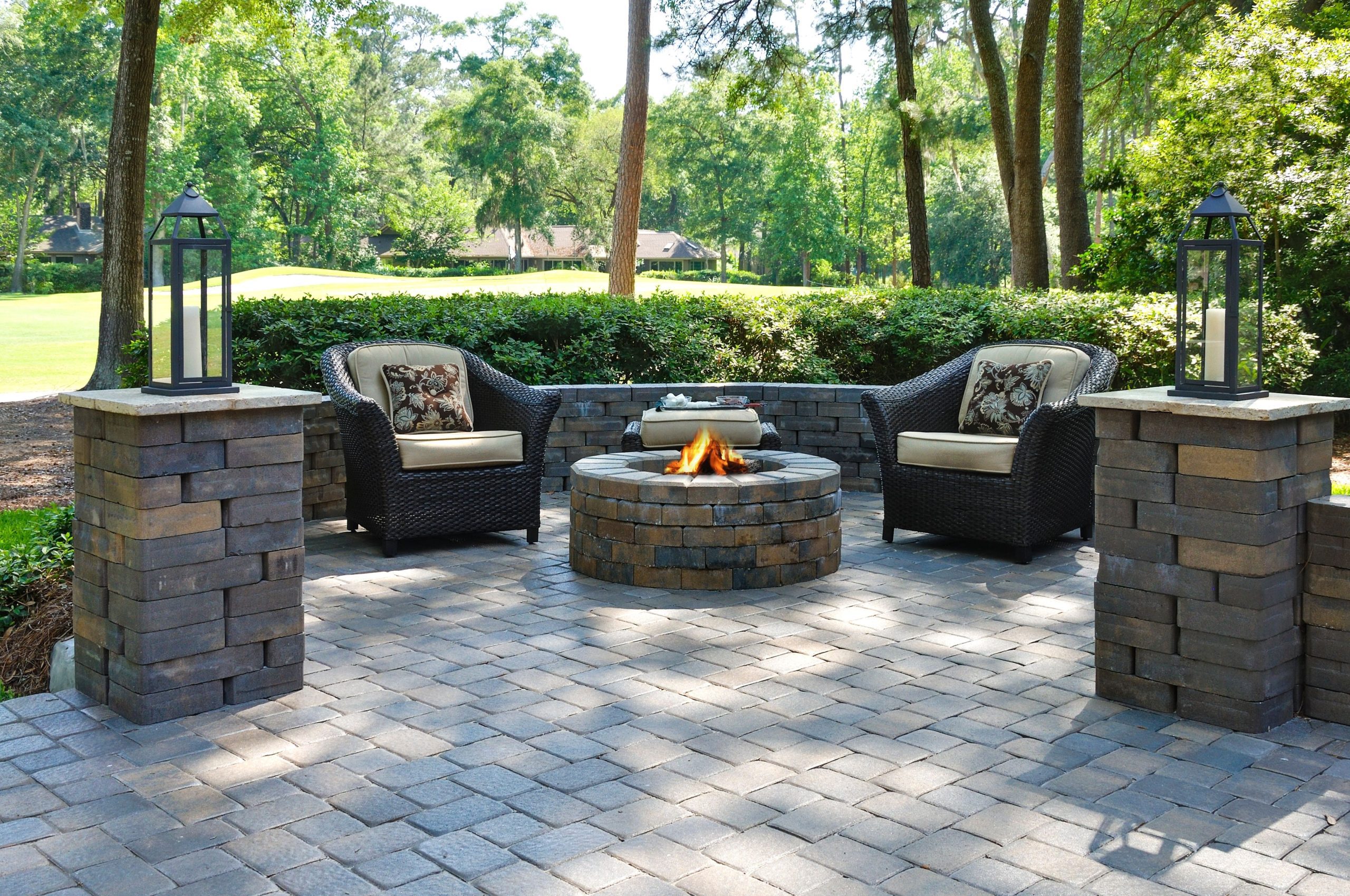 Two wicker chairs with cushions surround a stone fire pit on a paved patio, bordered by brick pillars with lanterns, set in a wooded backyard.