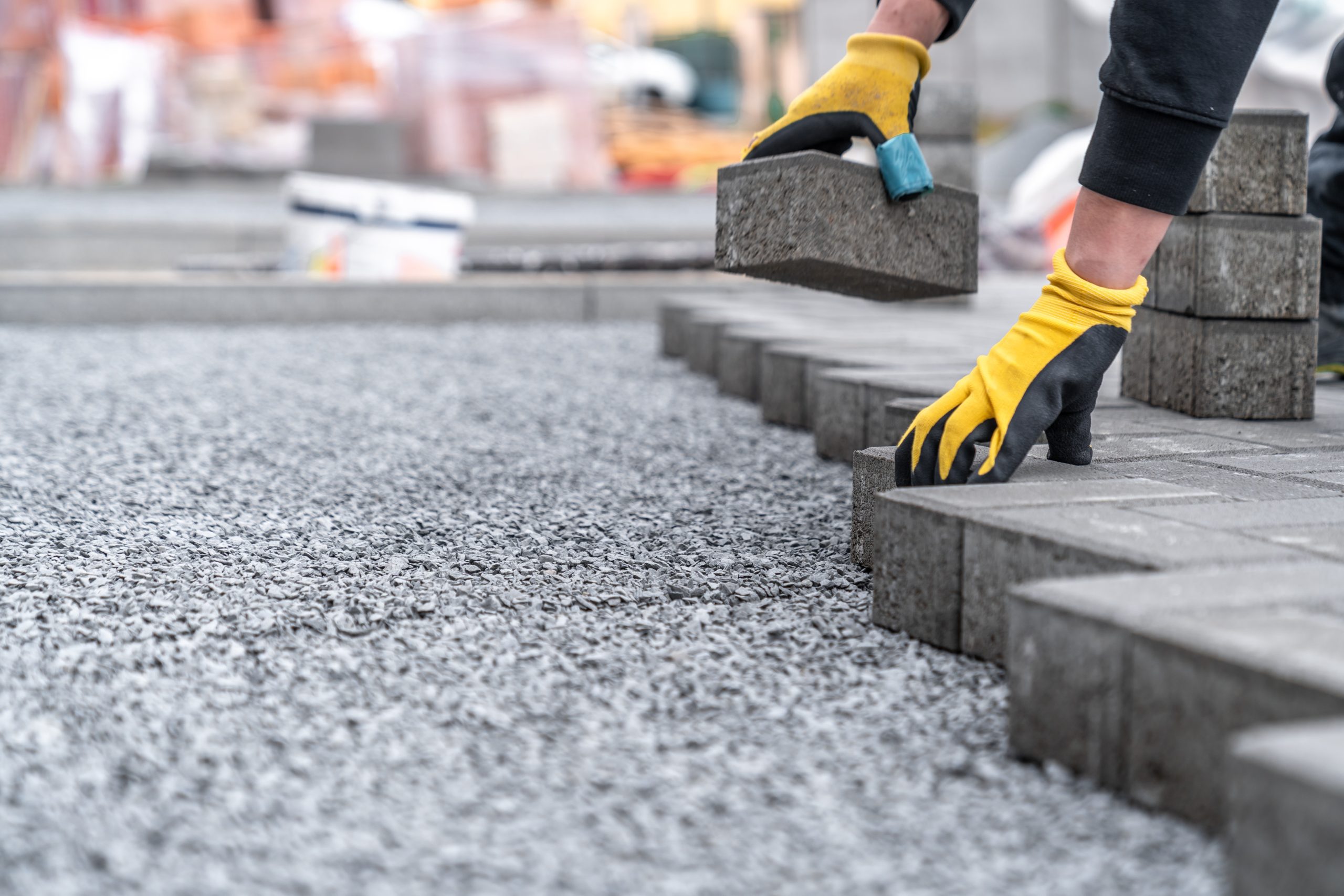 A person wearing yellow gloves lays concrete pavers in a row on a gravel surface, constructing a paved walkway or patio.