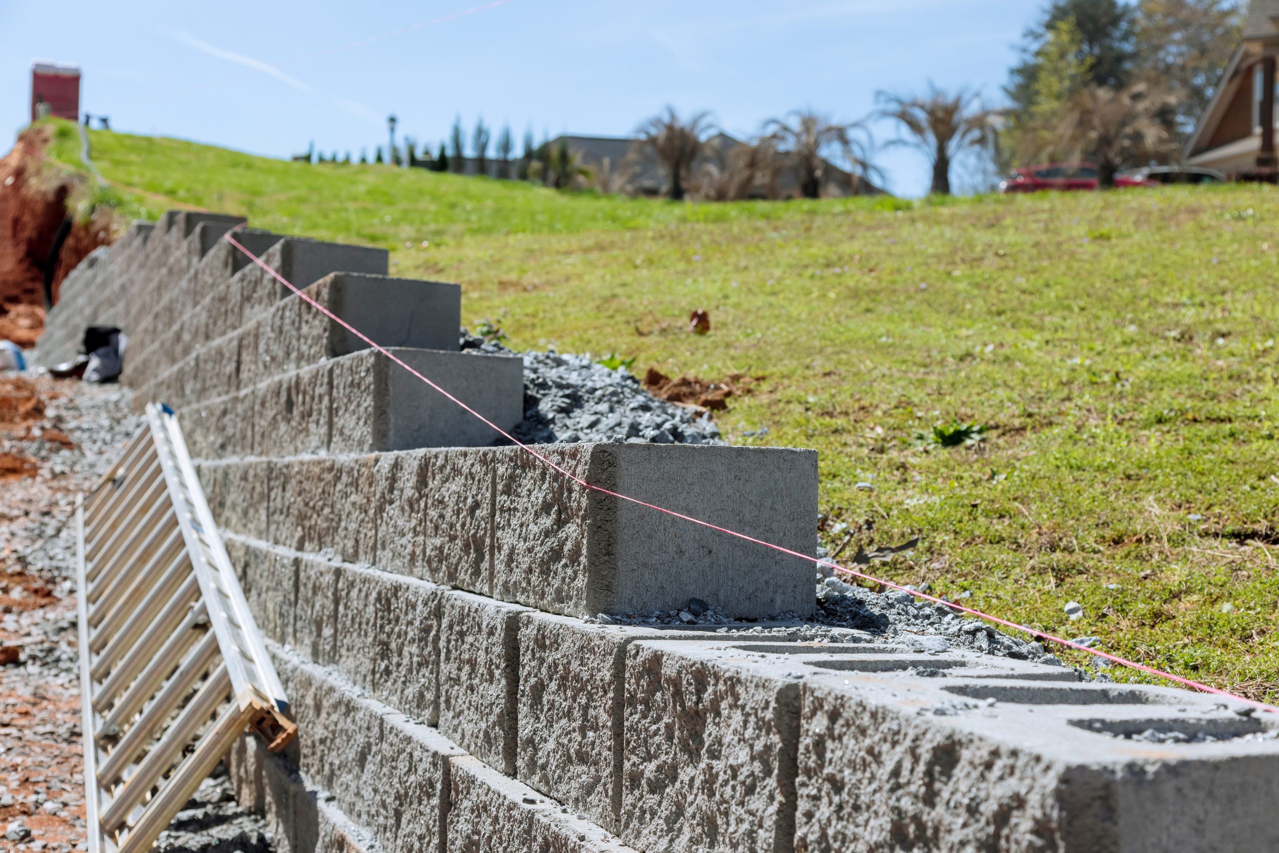 Partially constructed retaining wall with concrete blocks and string guides on a grassy slope; construction materials are visible alongside the wall.