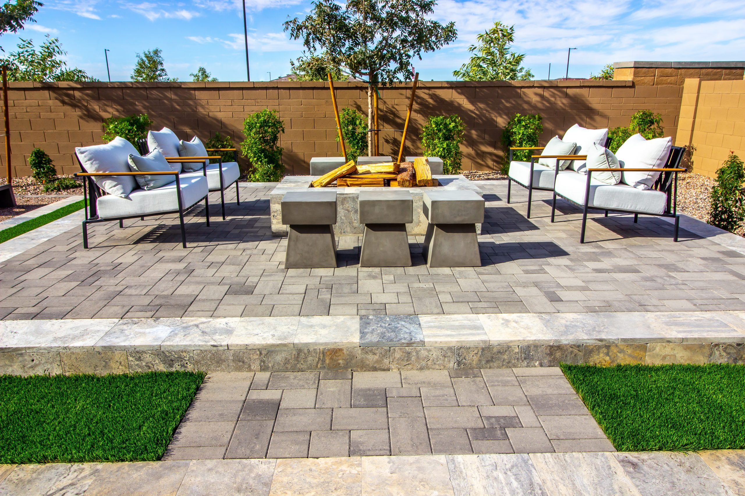 Modern outdoor patio with cushioned chairs, concrete stools, and a central fire pit on a paved surface, surrounded by greenery and a brown privacy wall.