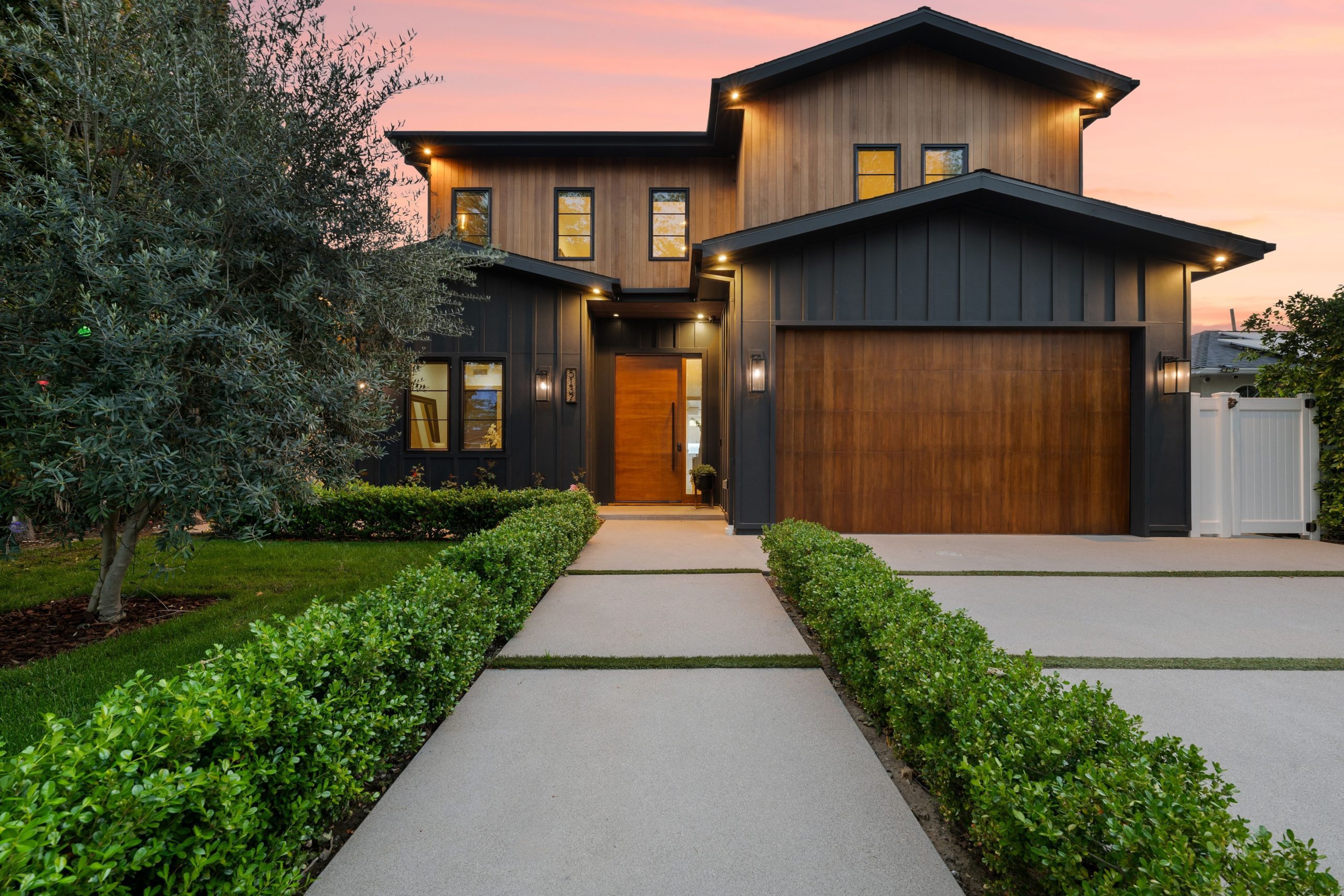 Modern two-story house with wood and dark panel exterior, large windows, attached garage, and landscaped front yard at sunset.