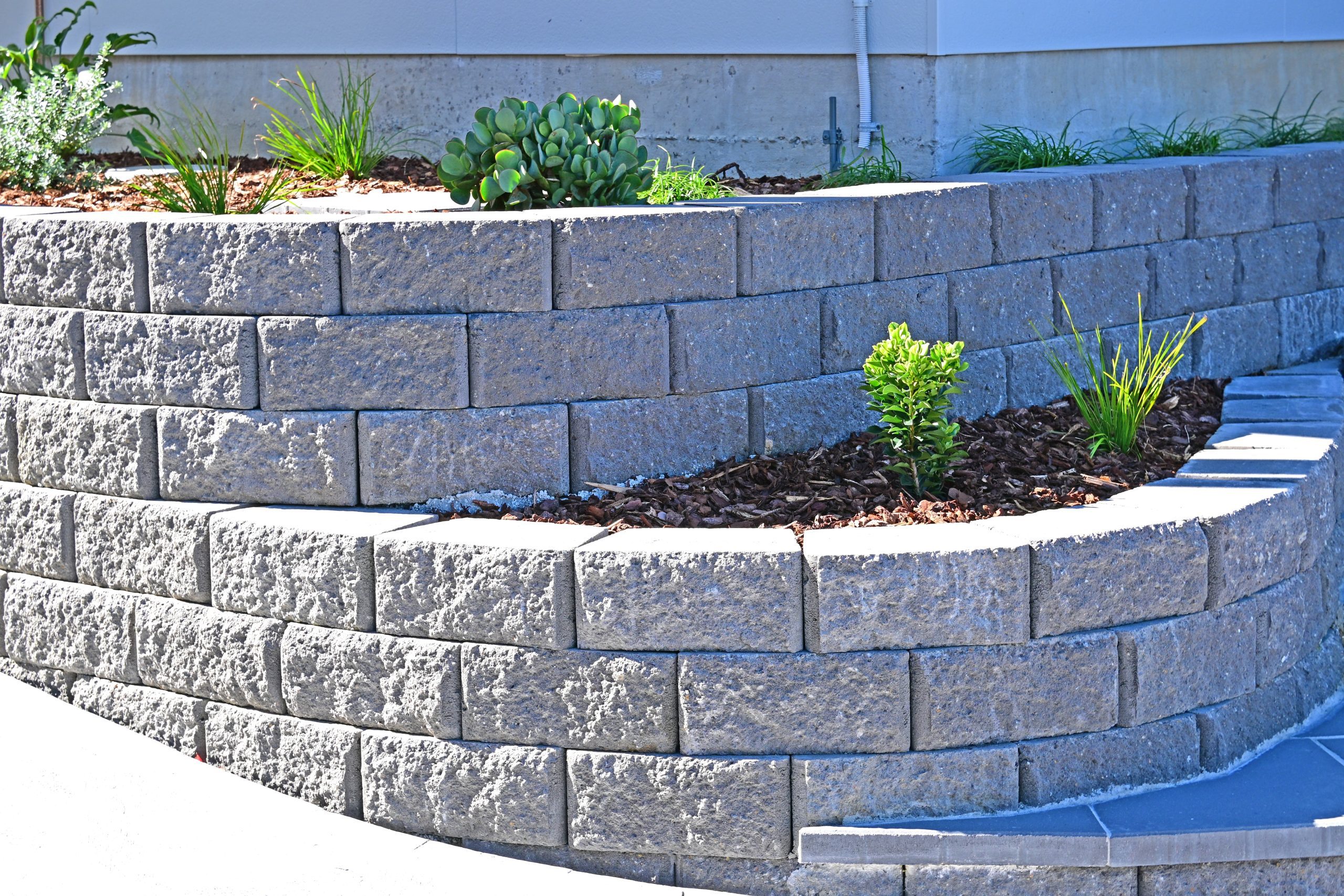 Tiered gray stone retaining wall with small green plants and mulch, located next to a building foundation.