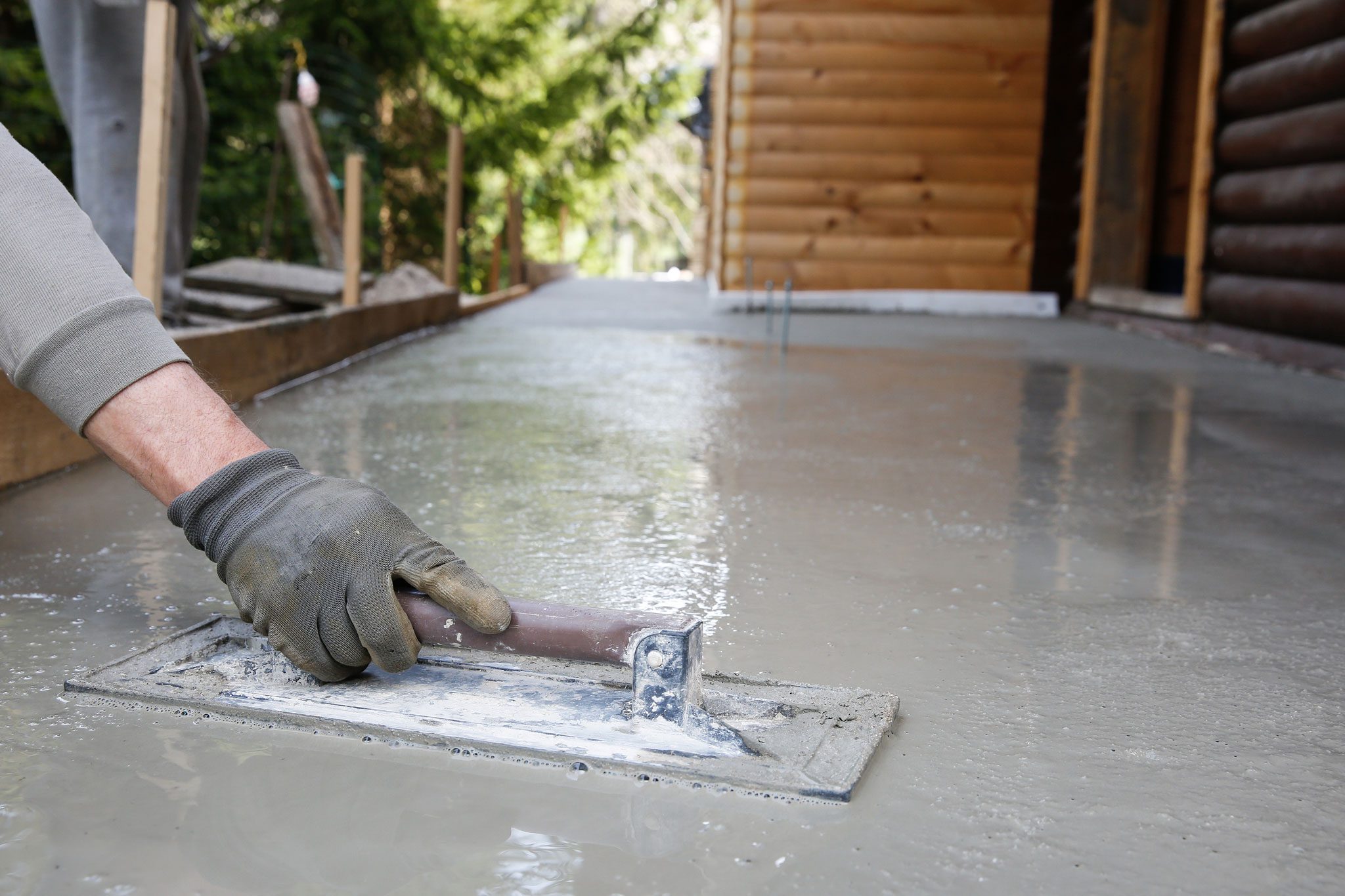 A person wearing a glove smooths wet concrete with a trowel outside a wooden building.