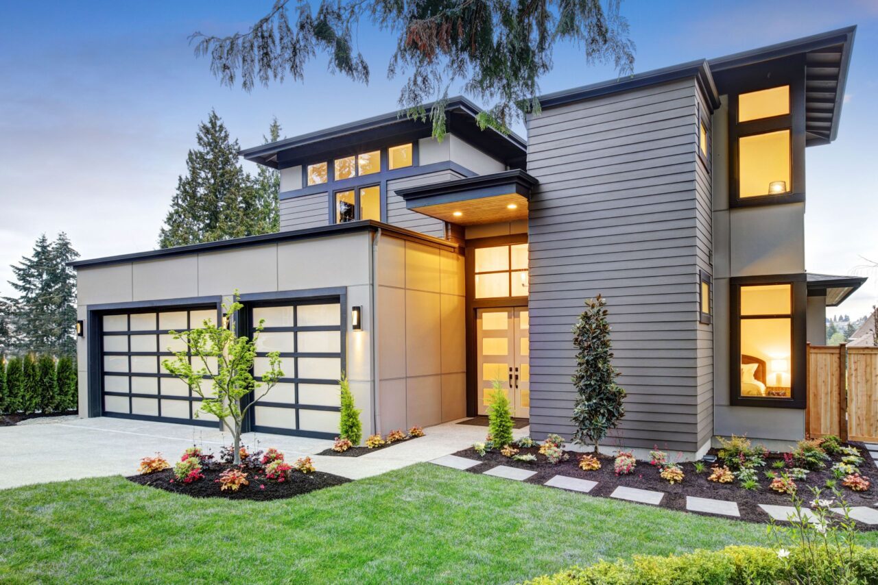 Modern two-story house with large windows, a double garage, and neatly landscaped front yard at dusk. Exterior features gray siding and geometric architectural lines.